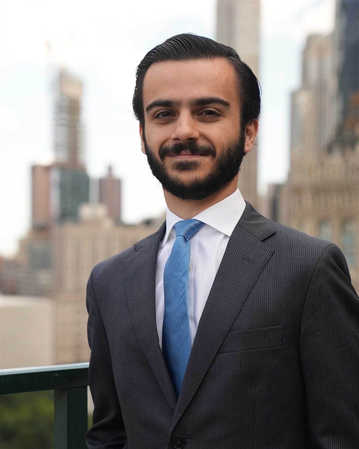 George P. Grimbilas in a pinstripe suit and blue tie, standing on a terrace with the Manhattan skyline behind him.